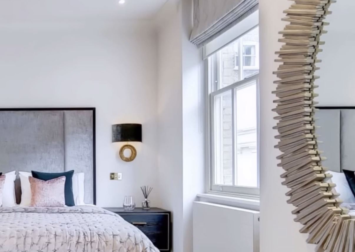 Bedroom with upholstered headboard, brass wall light and a sunburst mirror beside a sash window